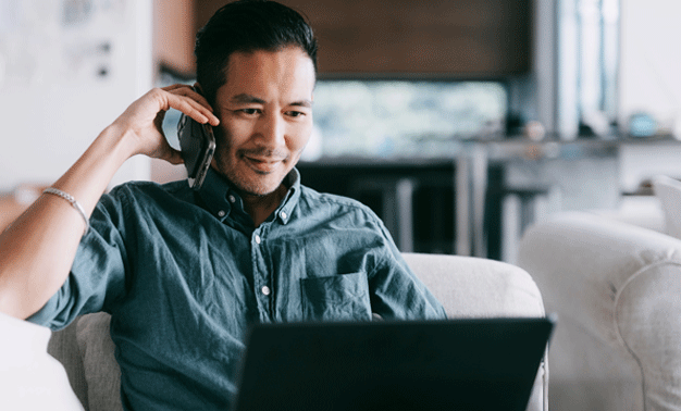 Man sitting at home, talking on a phone while using a laptop.