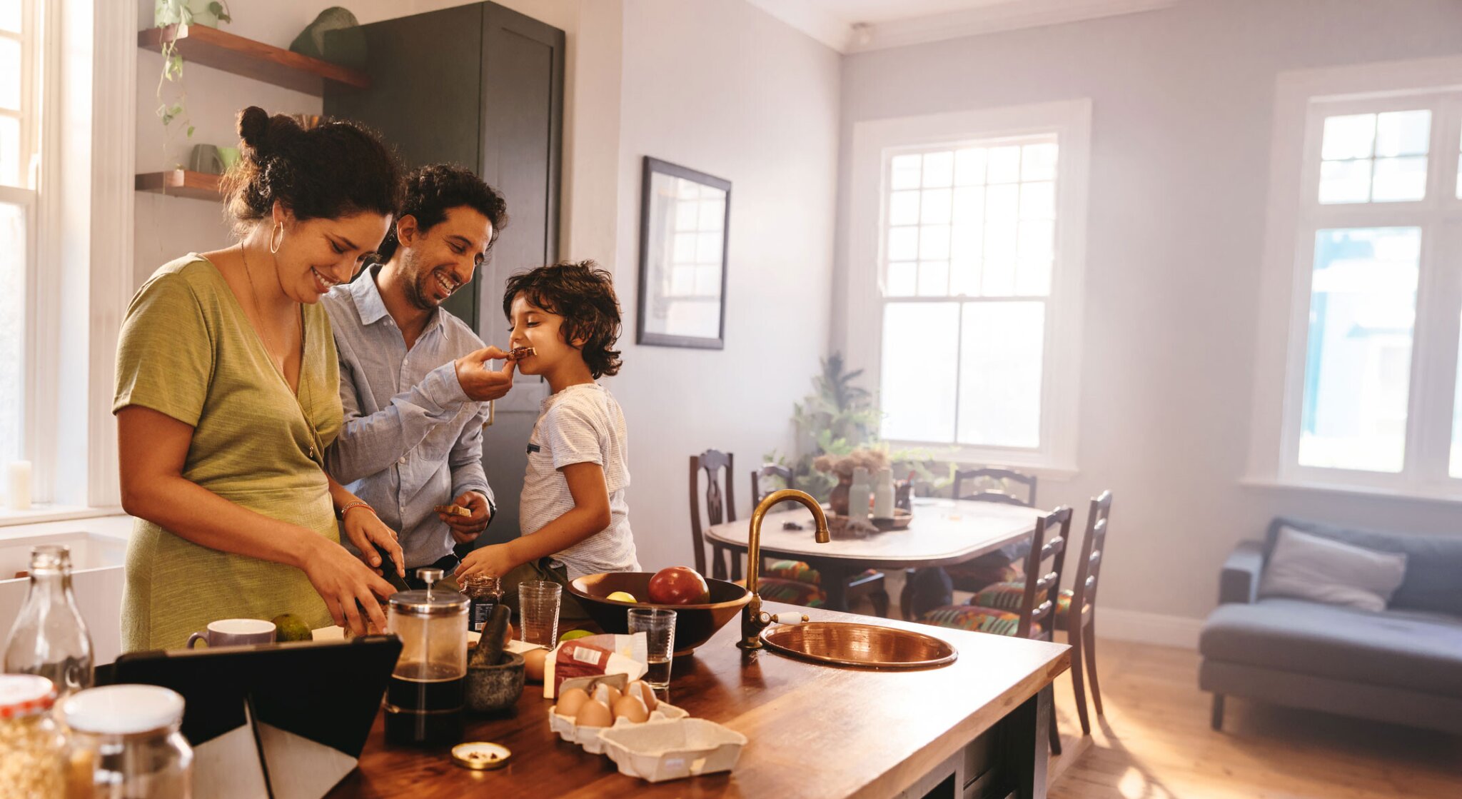 Playful dad feeding his son a slice of bread while his wife prepares breakfast.