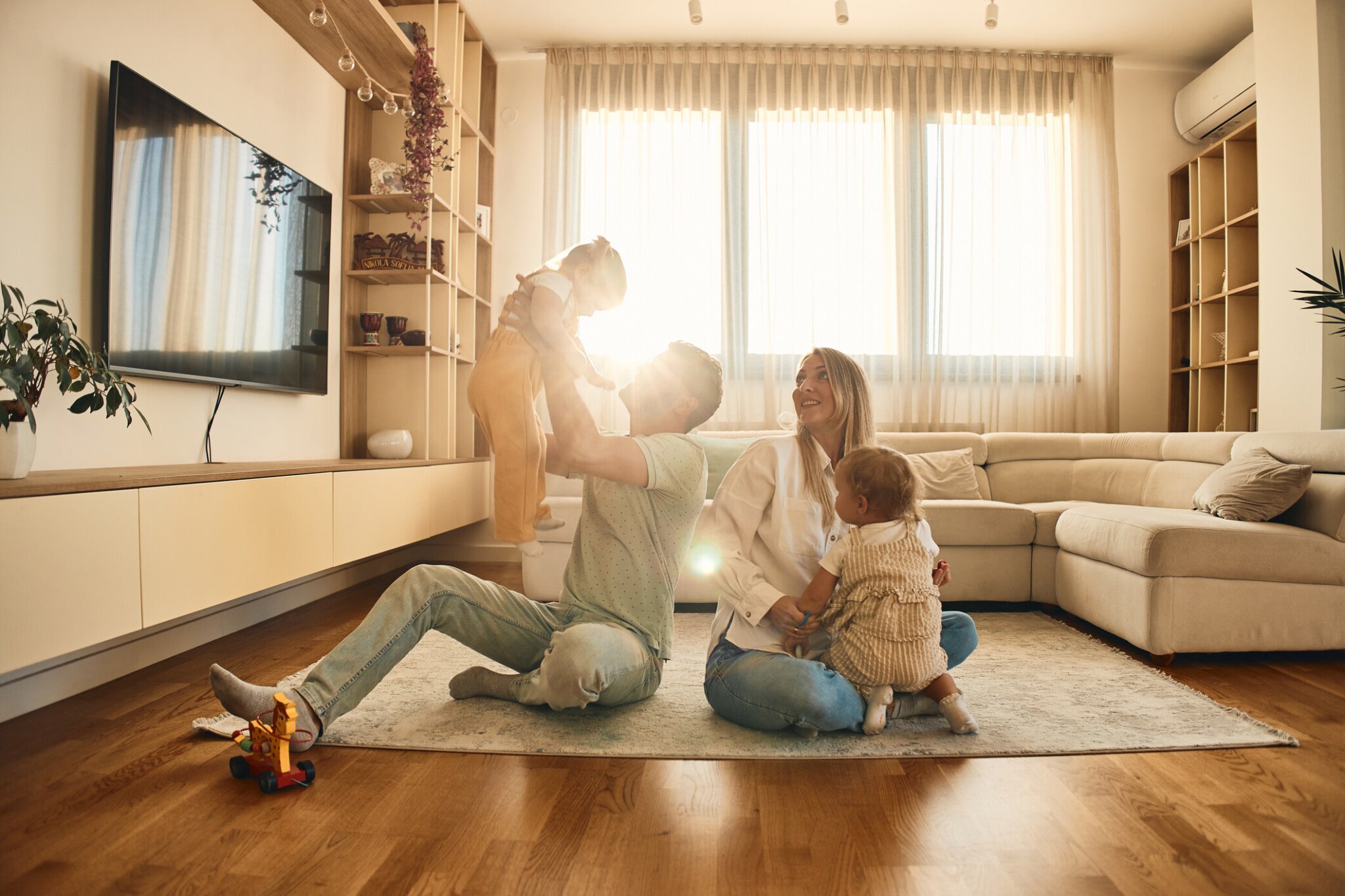 Parents sitting on a living room floor playing with their two young children in a sunlit home.