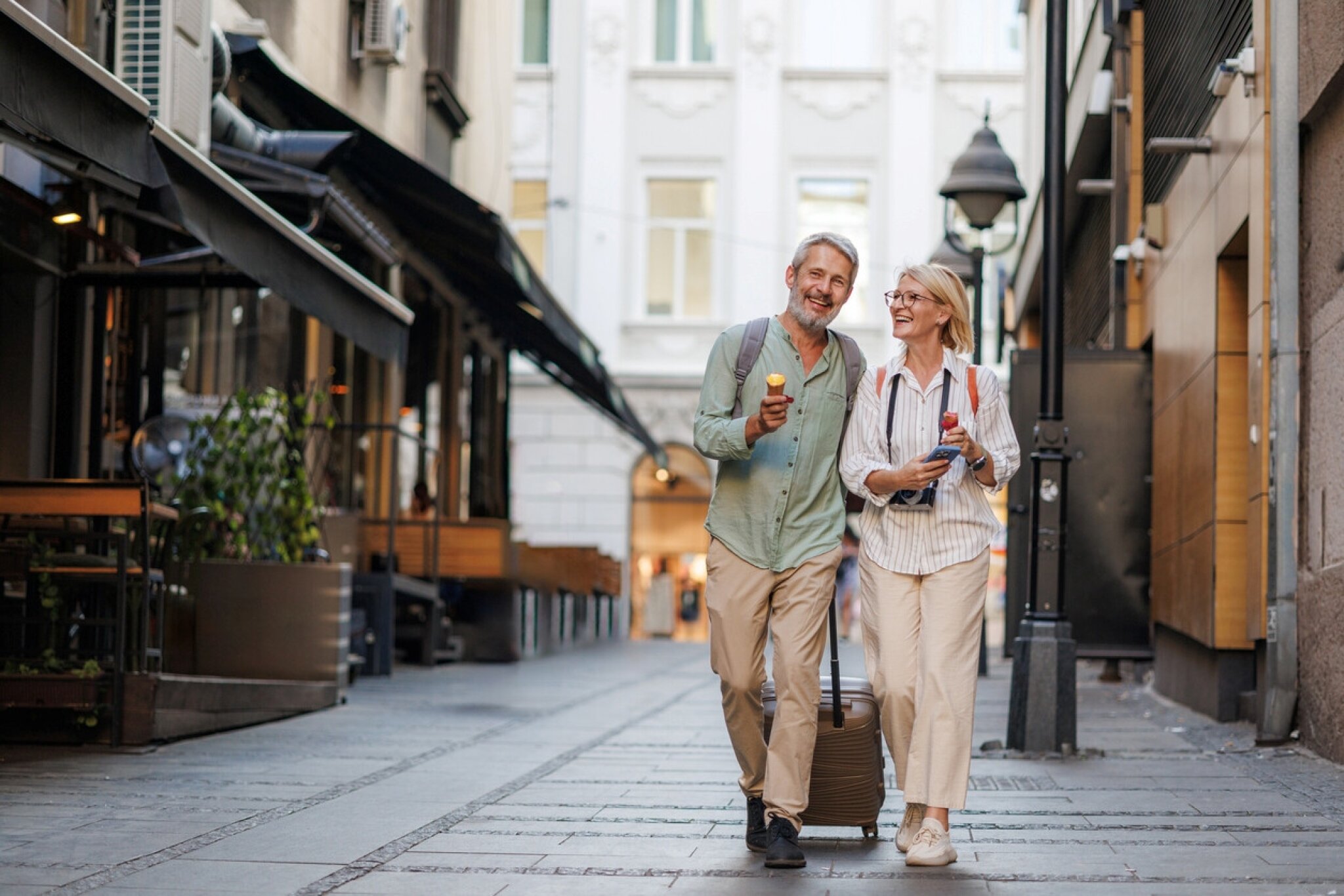 Older couple walking down a city street with a suitcase and holding ice cream cones.