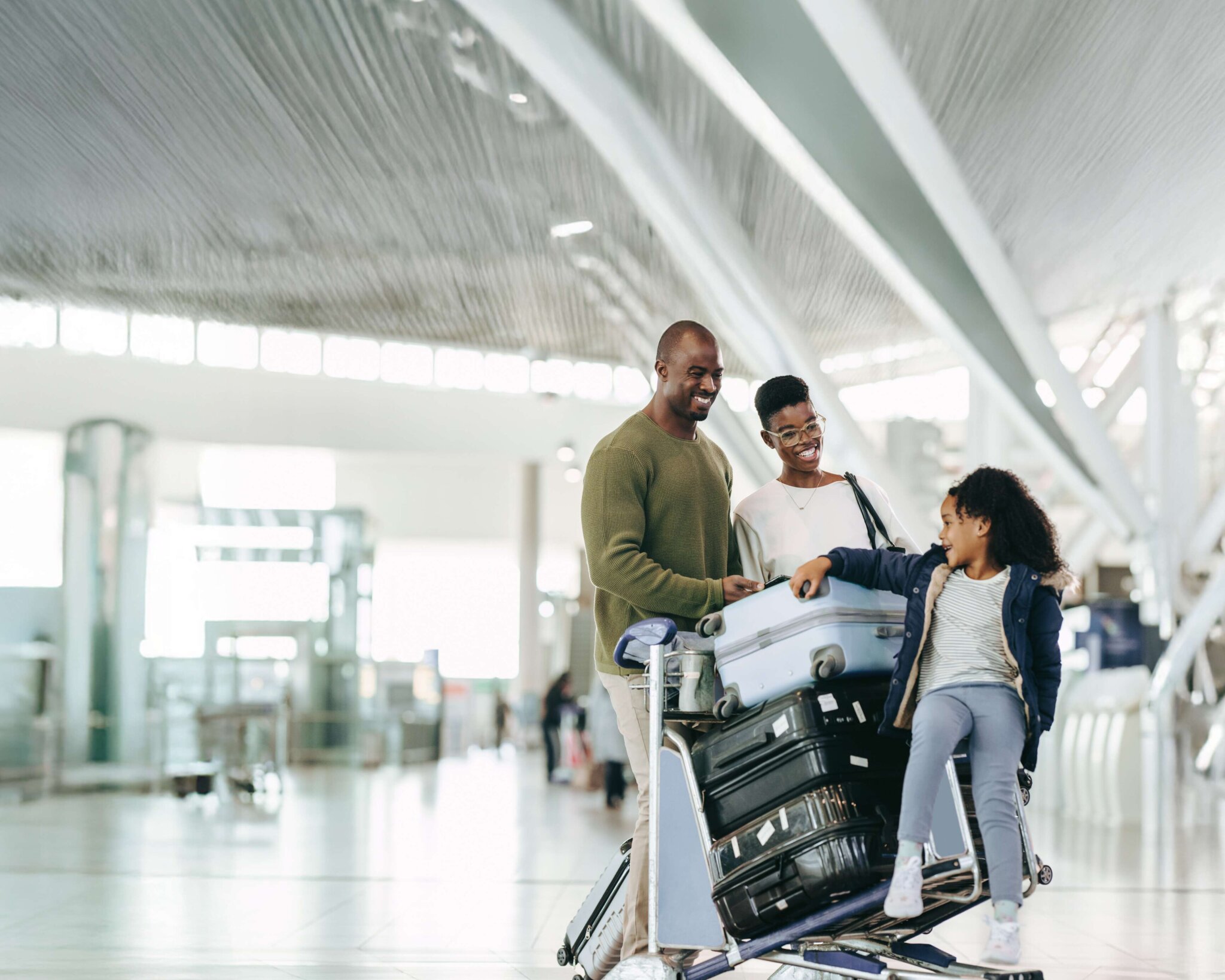 family smiling while walking through airport with daughter sitting on luggage cart.