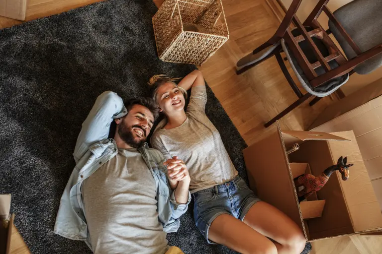 A couple holds hands while lying on their floor amongst packing boxes.