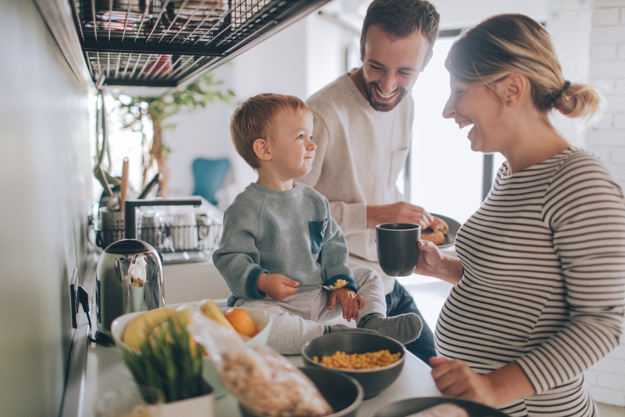 A family smiles and gathers around their eating toddler in a kitchen.