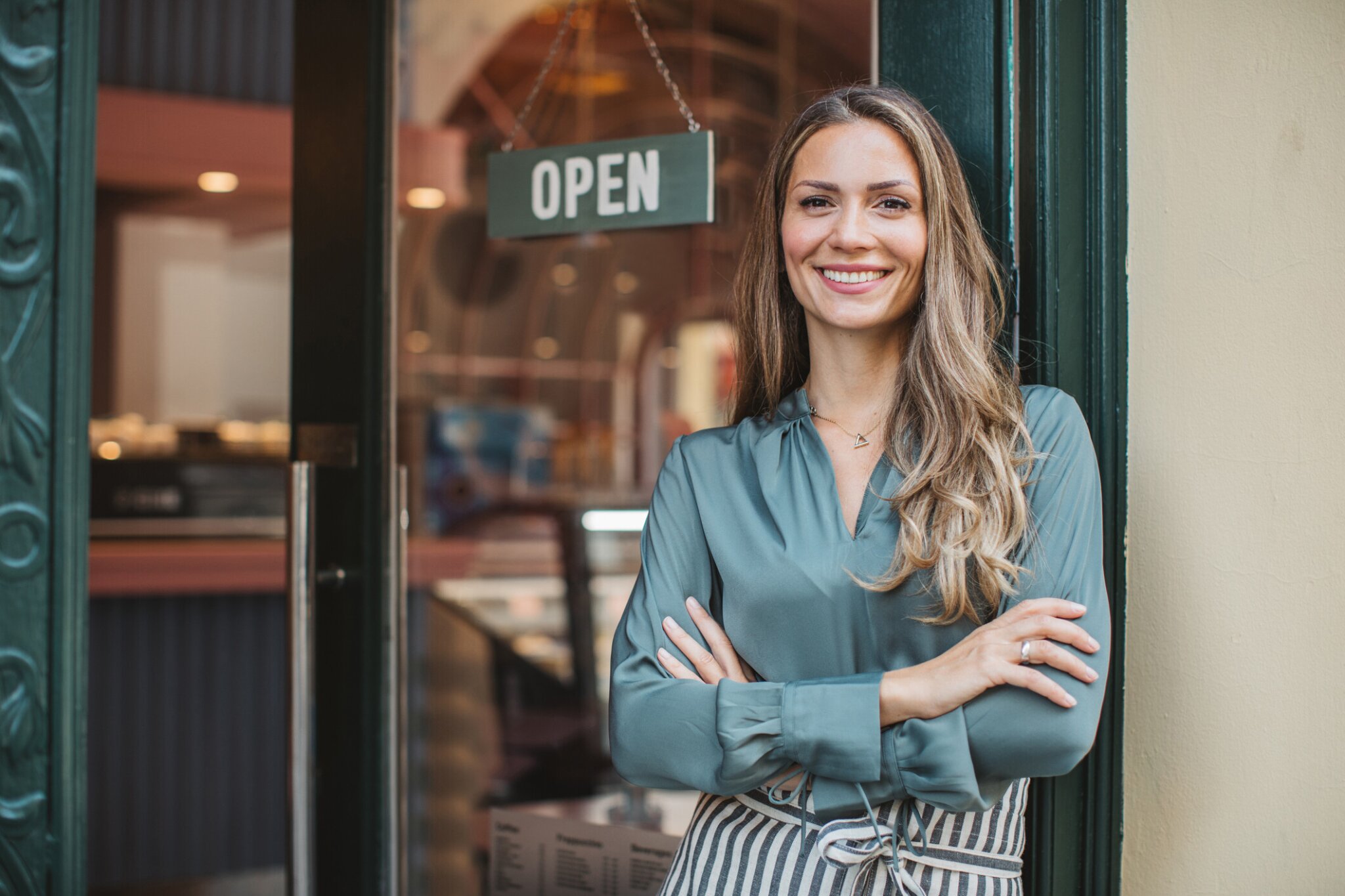 woman standing in front of open business with arms crossed smiling.