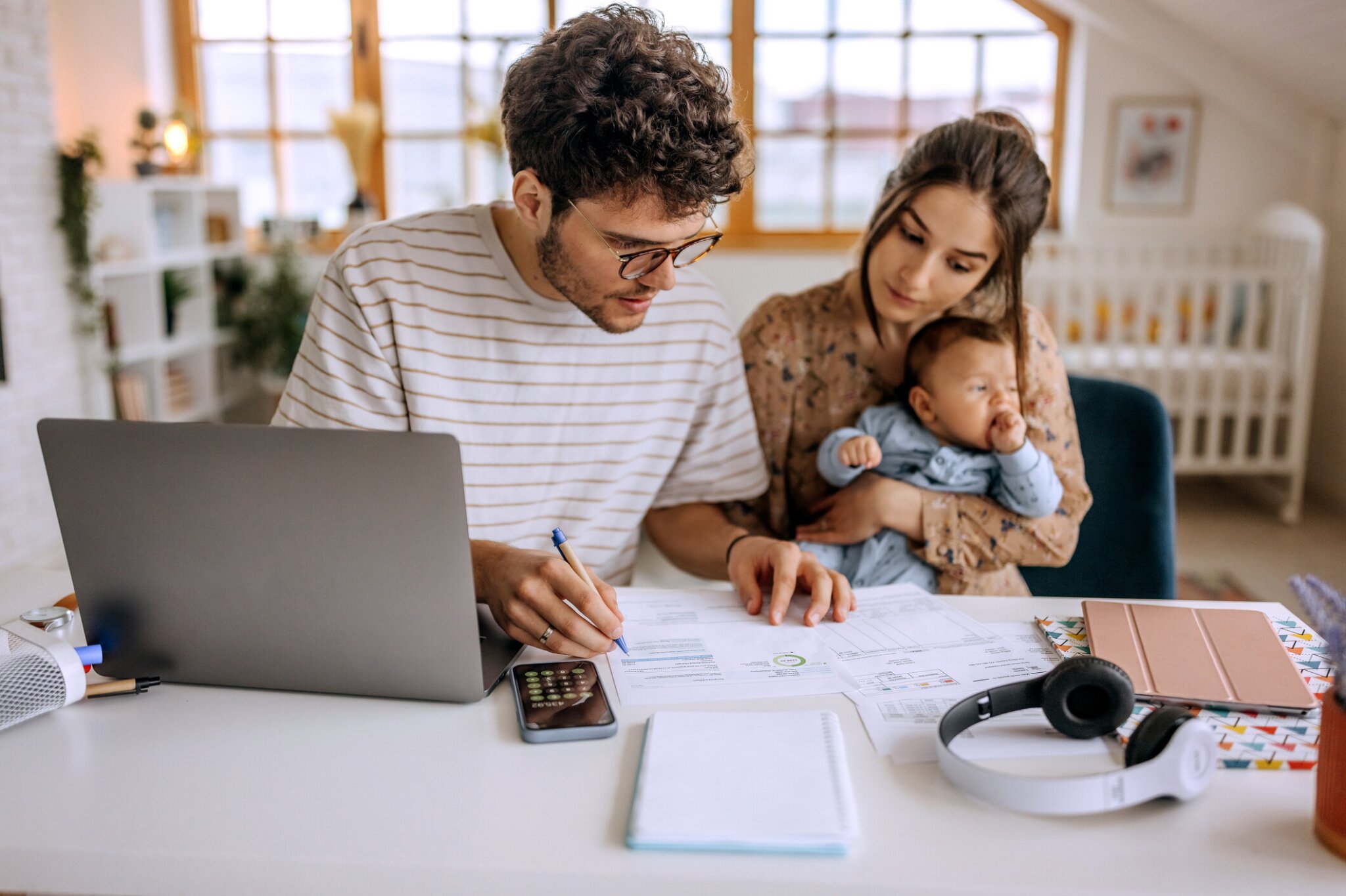 Man reviewing documents and writing at a table with a laptop while a woman holds a baby beside him.