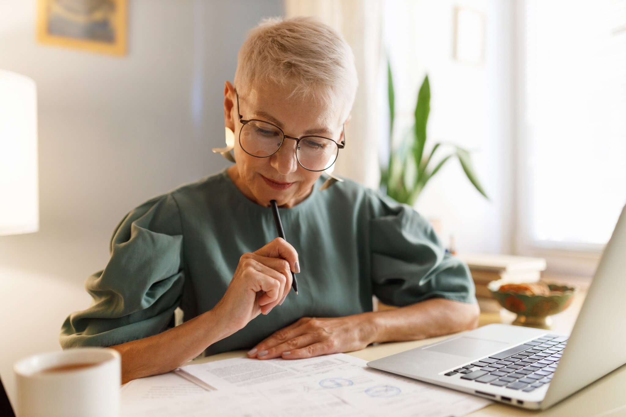 A woman sits at a table working with papers and a laptop.