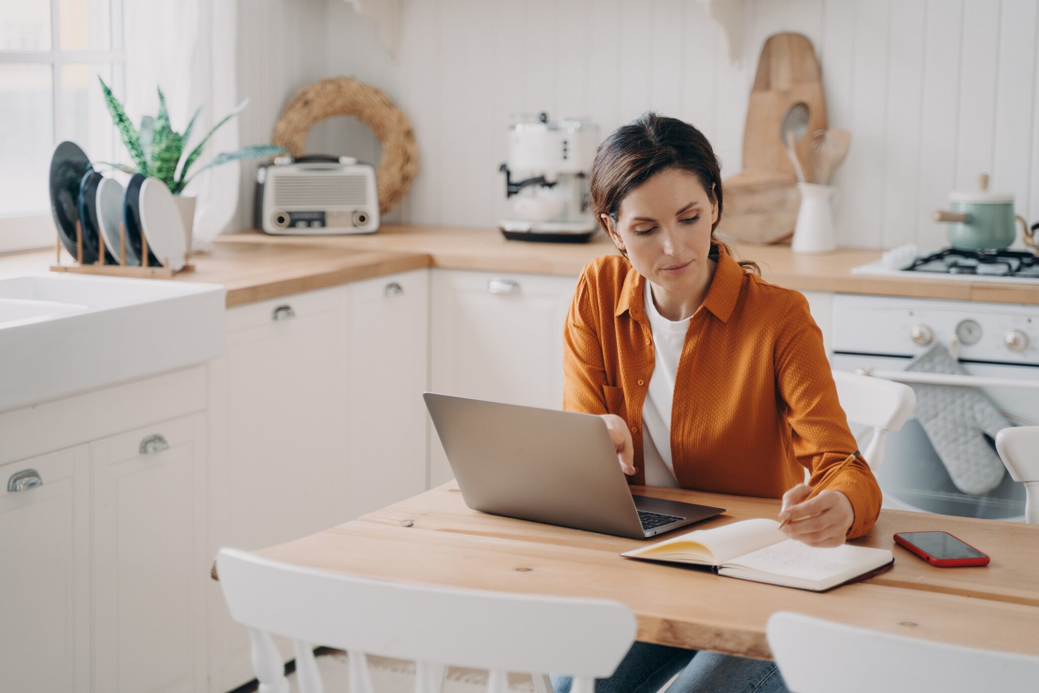 A woman works at her kitchen table with a laptop and notebook.