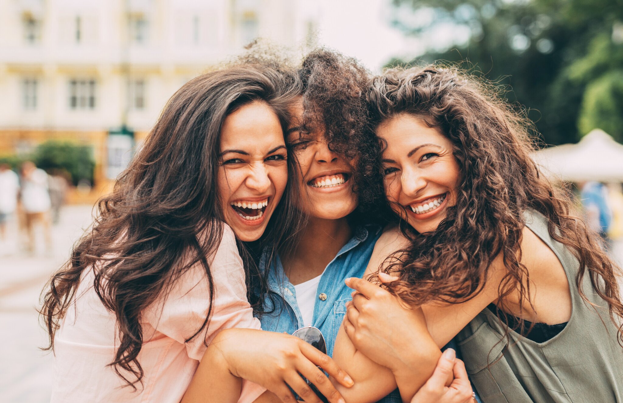 Three women smiling and hugging outdoors in a city setting.