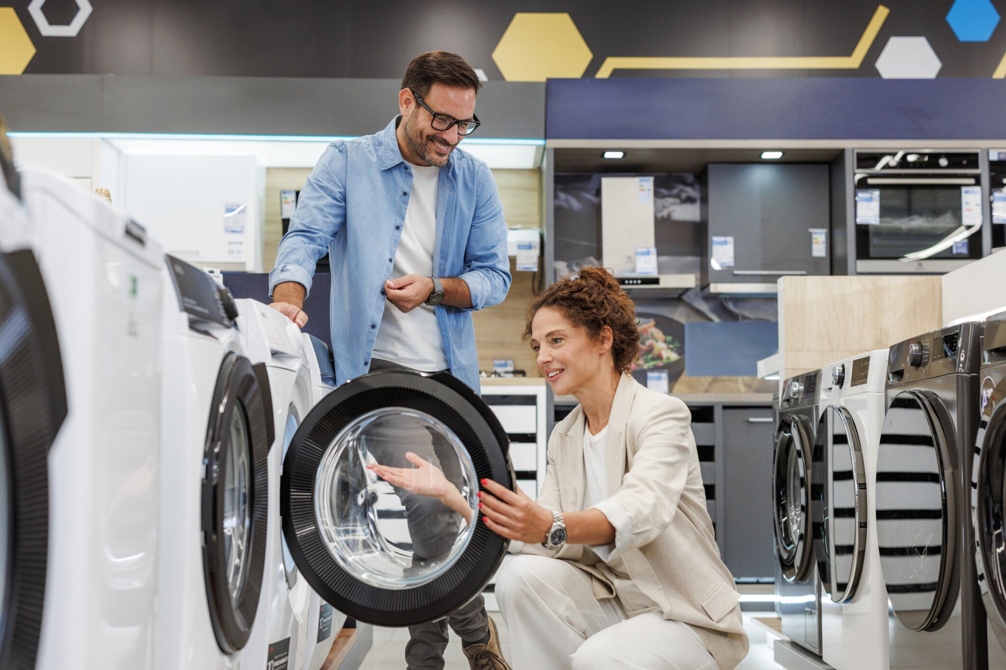 A couple shops for a washing machine in an appliance store, examining the drum of a front-loading washer while discussing options.