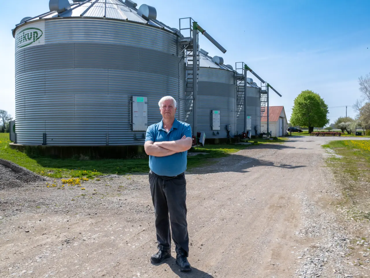 Man standing with arms crossed in front of large grain silos on a farm.