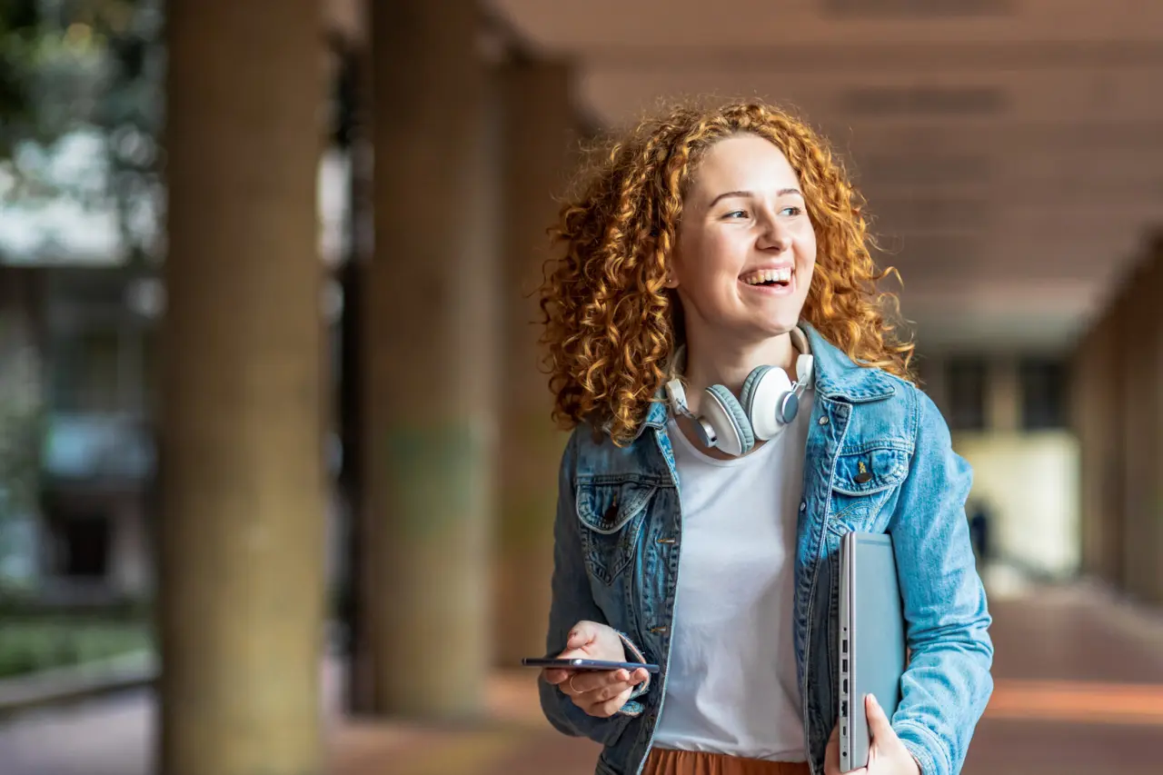 Young woman smiling holding phone and computer while walking through college campus.